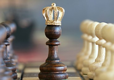 A pawn sits in the middle of a chessboard with a crown on it. Other pawns of white and brown are on either side.