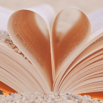 An open book with its centre pages curled together to form a heart. The book sits in a pile of pebbles and sand, and there is sand on one of the pages too.