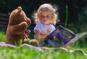 Little girl reading a book to her teddy bear on a picnic blanket on grass