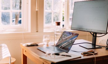 A desk in front of a window containing a large computer monitor and a laptop, as well as a phone ,a class of water, a pen, a notepad, and glasses.
