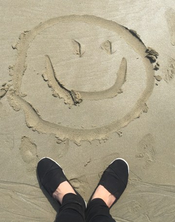 A smiley face drawn in the sand that is framed by a woman wearing black shoes and pants