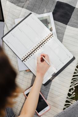 Woman sitting on a picnic blanket and writing in a weekly planner with her phone close by