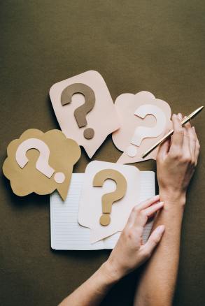 Construction paper question marks on different colored backgrounds and shapes.  All lead to a notebook and a woman's hands which hold a pen