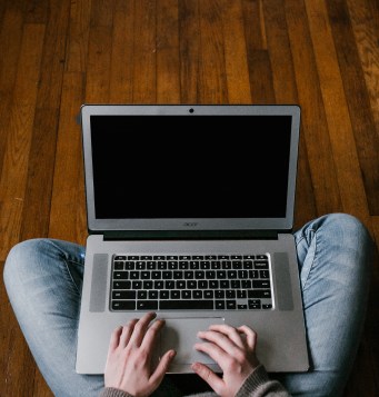 Person wearing jeans and a sweater sitting cross-legged on the floor with a laptop on their lap.