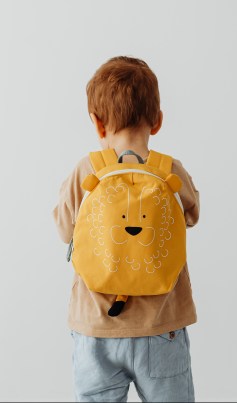 A red-headed toddler with facing away fro the camera in a brown shirt and jeans. They are wearing a lion shaped backpack.