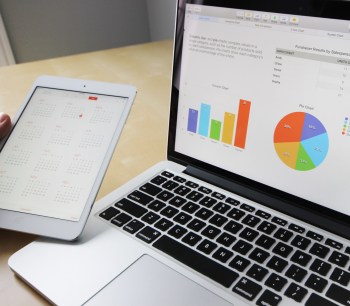 A laptop sits open on a statistics page and someone is holding a calendar open on a tablet