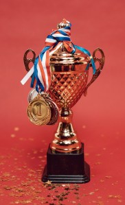 With a red background, a gold trophy is wrapped with 1st, 2nd, and 3rd place medals on their ribbons. There is gold star confetti spread around.