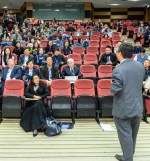 A male professor lectures in an auditorium with a crowd of students in red chairs