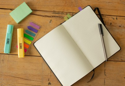On a wooden desk is a notebook with sticky notes stuck in its pages and a pen on top. Next to the notebook are highlighters and more sticky notes.