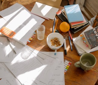 A round table is covered in open study books, a calculator and pens, class notes, sticky notes, and more textbooks. There is also an empty glass, a tea mug, and a bowl of snacks.