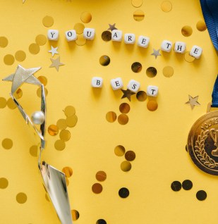 A yellow table has round golden confetti, a medal, and a trophy. There are white beads with black letters saying "you are the best."