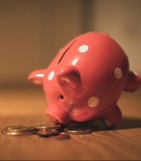 A white spotted pink piggy bank has its nose down in a small pile of coins on a table.