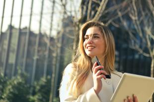 A business woman stands in the sun outside a gate. She wears a white coat and holds a smart phone and tablet in her hands.