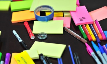 There are permanent markers, pens, highlighters, a roll of blue tape, a compass, pencils, and various sizes and kinds of sticky note on a black table.