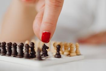 A woman with red-painted fingernails moves a black knight on a mini chessboard