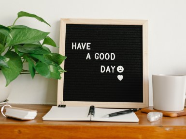 Scene focuses on a wooden desk. There is a felt letter board reading "Have A Good Day" with a smiley face and a heart. Next to that is a leafy plant in a white planter. Theer is a flip card book and next to that is an open notebook with a binder clip and a black pen. Next to that is correctiont ape and a cup of coffee on a wooden coaster.
