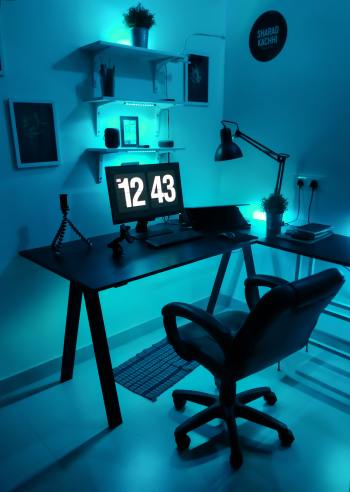 A modern work-from-home set up featuring a black flat screen computer monitor on black wooden table. The computer screen reads the time 12:43. There are decorative shelves on the wall with odds and ends on them. The entire space has blue mood lighting.