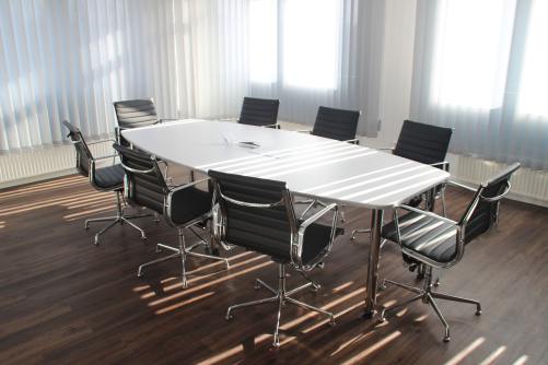 A white board room table with 8 black rolling chairs sits in an office room with white blinds