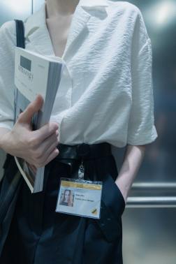 A woman dressed in a white blouse and black pants stands in an elevator. She has a textbook, a shoulder bag, and a name tag with her photo on it.