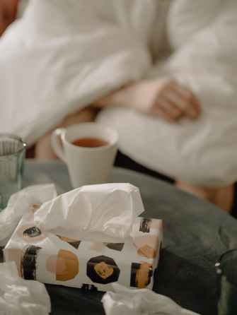 A cup of tea sits on a coffee table with a box of tissues and wadded up used tissues. In the background there is a person wrapped in a duvet.