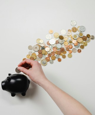 A person puts a coin in little black piggy bank on its side. There is a cloud of coins on the tabletop that gives the appearance of steam from a train.