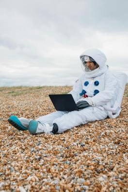 An astronaut uses a computer while sitting on a pebbled beach.