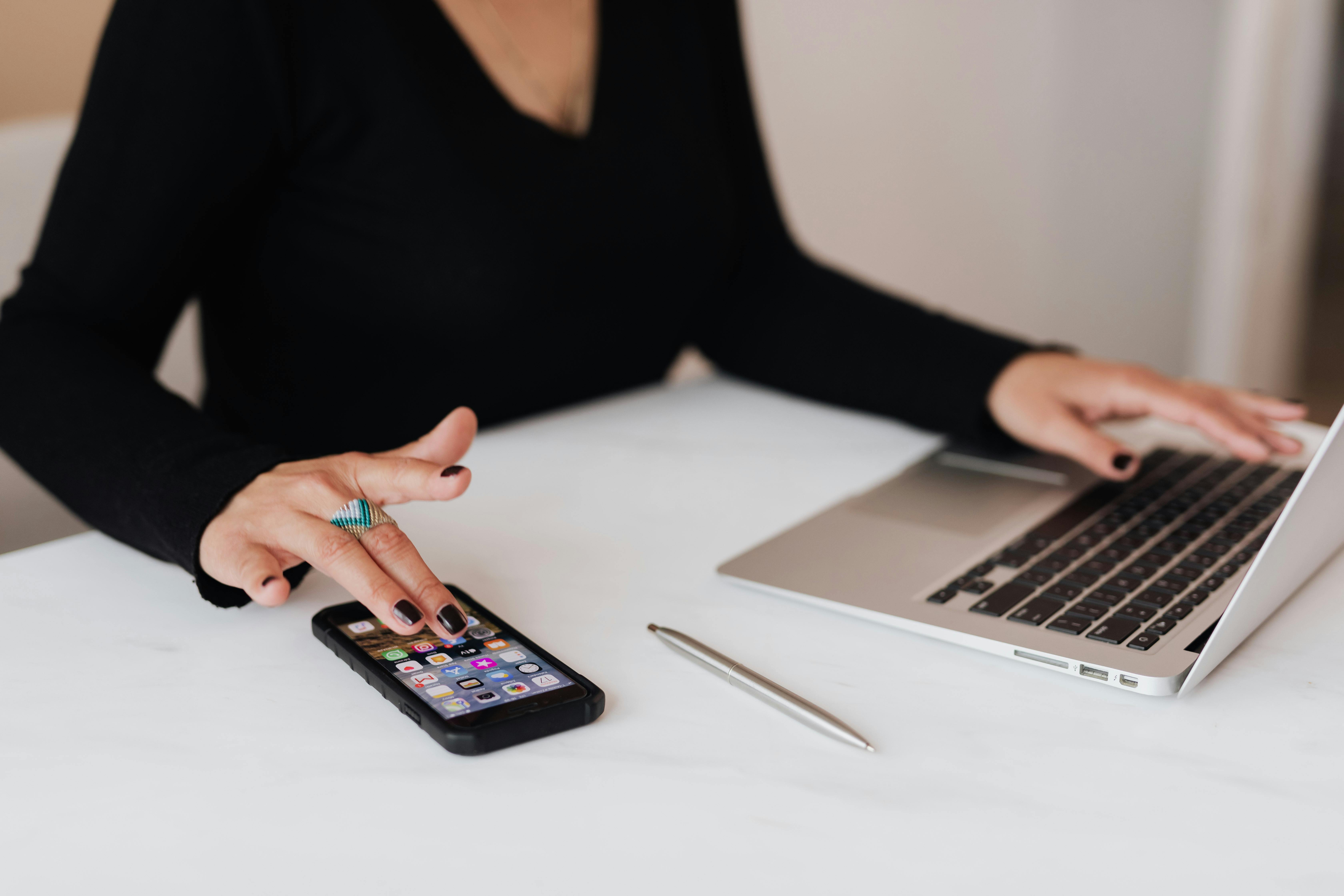 A woman with a black shirt and black fingernails sits at a white desk. She is using a computer with one hand and accessing her smartphone with the other.
