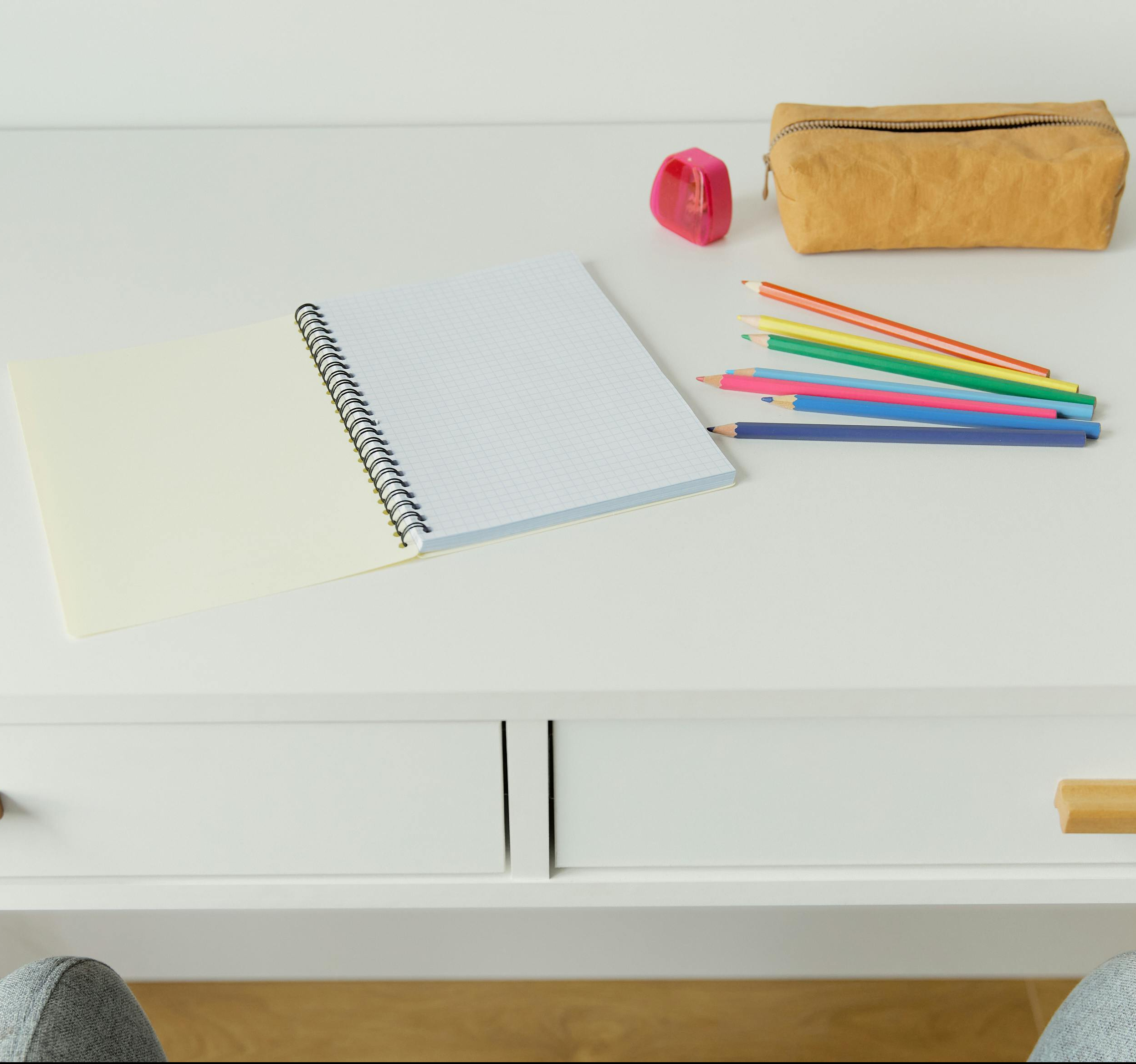 A blank, spiral-bound notebook sits on a wide desk. Next to it sits coloured pencils, a sharpener, and a leather pencil case. In front of the desk is a heathered grey office chair.