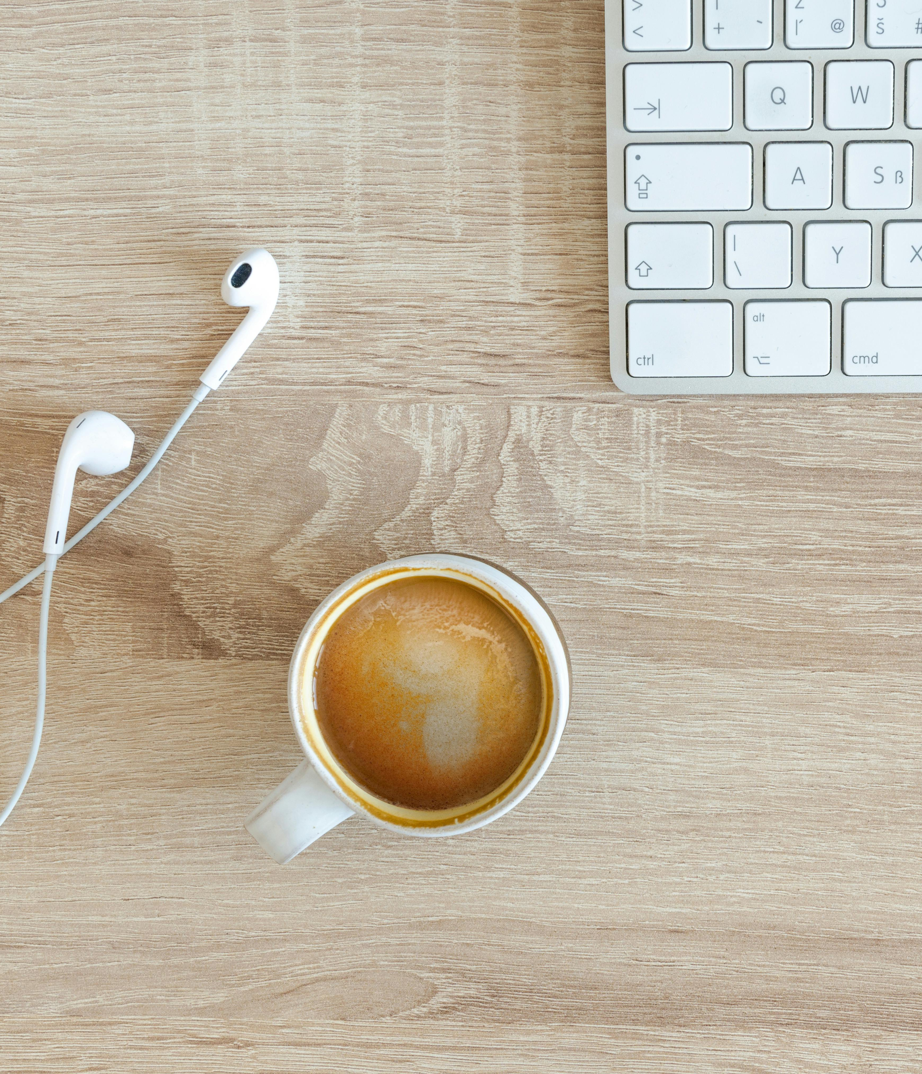 A top down photo of an iMac keyboard sitting on a wooden desk with earbuds and a small cup of coffee nearby.
