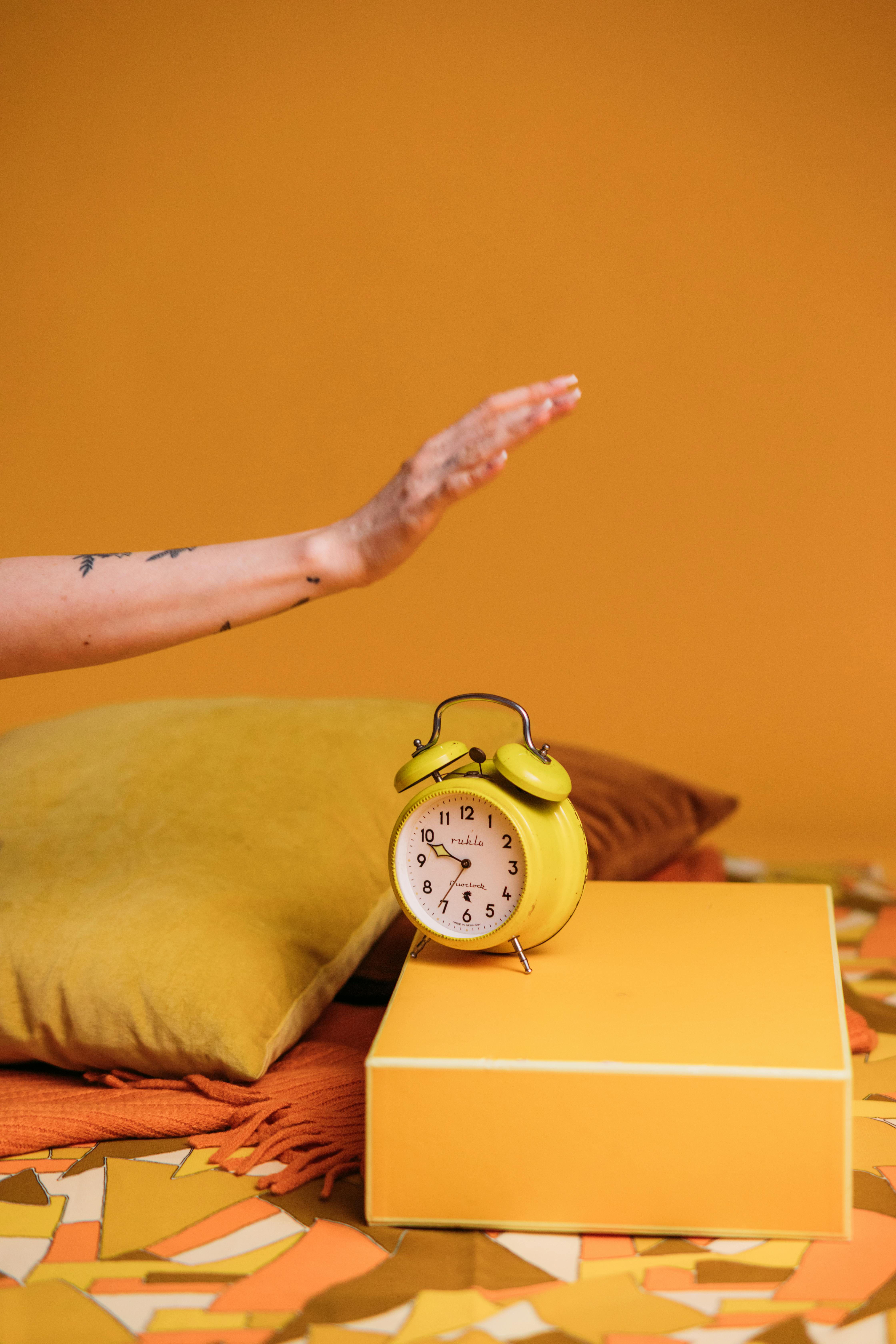 A yellow old-fashioned alarm clock sits on a yellow platform surrounded by yellow and orange pillows. There is an orange wall and '70s-style pattern flooring. A tattooed woman is reaching out from the left of the photo to turn the alarm clock off.