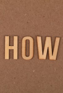 Wooden cutout letters read "HOW" on a plain cardboard background.