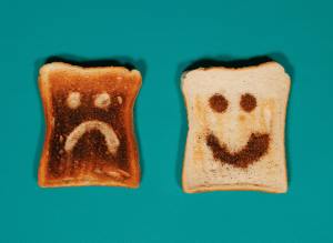 Two pieces of toast sit on a green surface. On the left is a burnt piece of toast with a sad face on it. On the right is a golden piece of toast with a happy face on it.