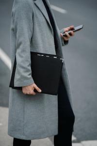A woman walks with a business portfolio in one hand and a smartphone in the other. The woman wears a grey, wool pea coat.