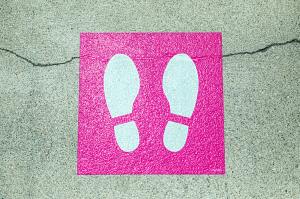 On green concrete, a pink square is painted with white bootprints.  Indicates how to stand in line.