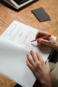 A woman is editing a script with a red marker. She has pink nail polish and is working at a weathered wooden desk. To her right is a closed black notebook.