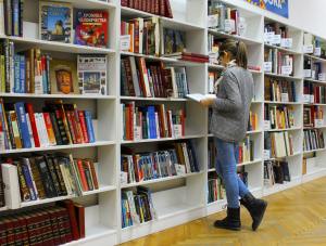 A woman stands in front of a shelf of books in a library. There are organized shelves on both sides of her. She is reading a book. She is wearing jeans, a grey sweater and a floral scarf. She has glasses and brown mid-length hair pulled back into a ponytail.
