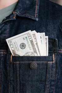 An up-close photo of person wearing a jean jacket. In its pocket are a selection of USD bills sticking out
