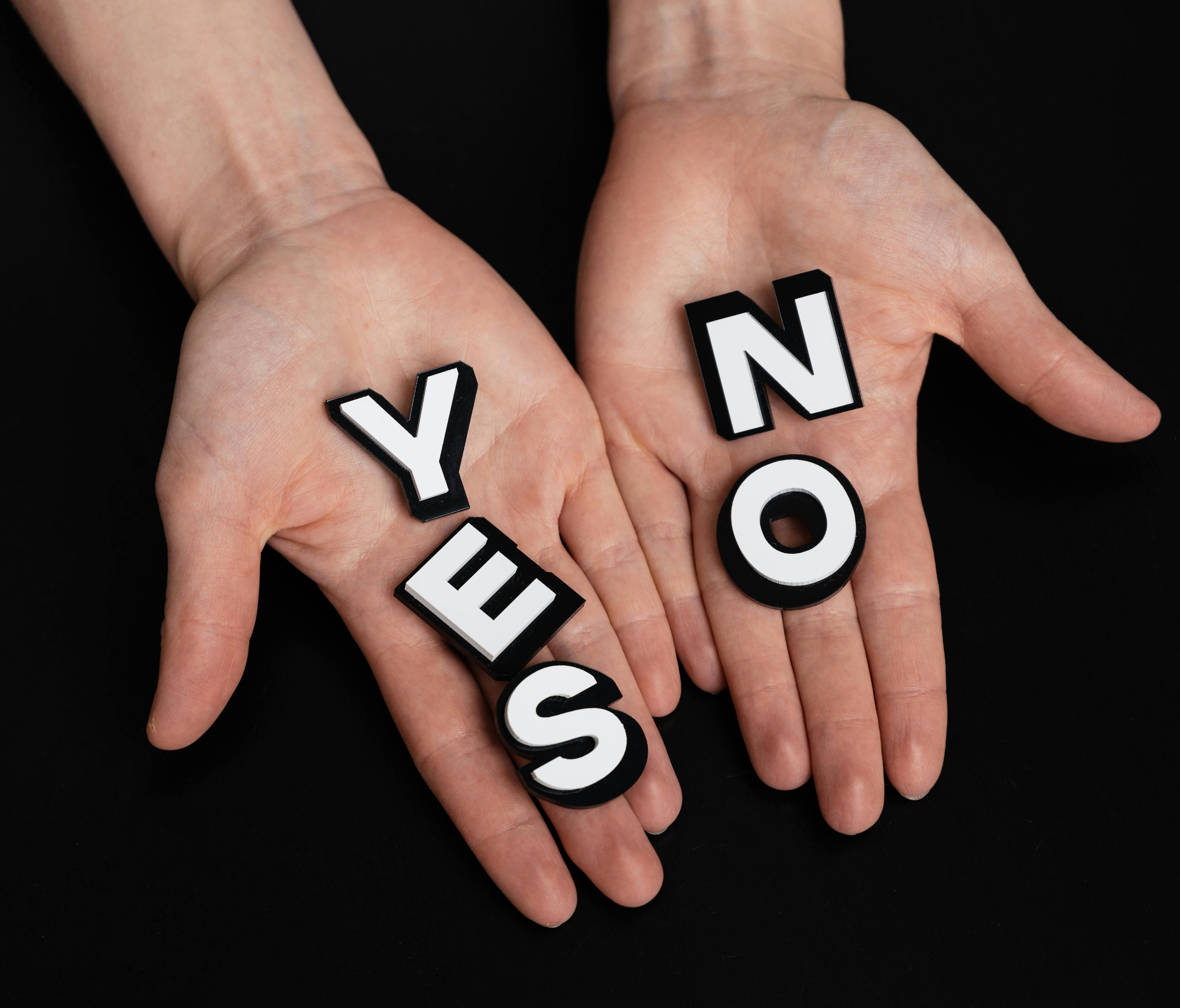 Two hands are facing palm up against a black background.  In one hand is spelled out "YES" in white and black 3D effect stickers.  In the other hand is spelled out "NO" in black and white 3D effect letters.