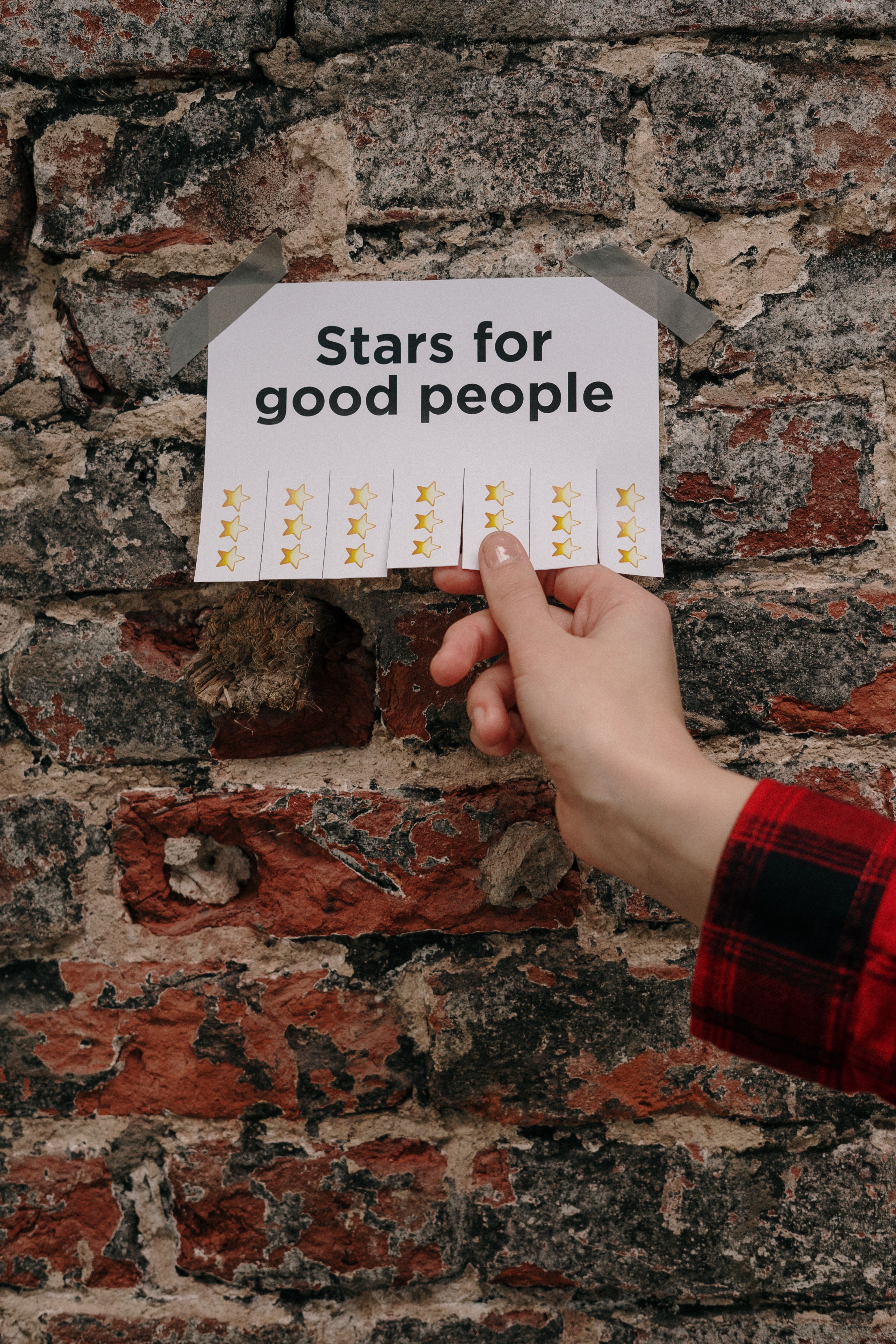 A piece of paper taped to a distress brick wall reads "Stars for good people". There are tabs you can rip off with gold stars on them. A person wearing a red and black plaid shirt is in the process of pulling a tab off.